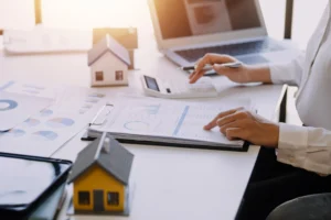 Profissional analisando relatórios e indicadores de gestão imobiliária em escritório com calculadora, notebook e miniaturas de imóveis sobre a mesa.